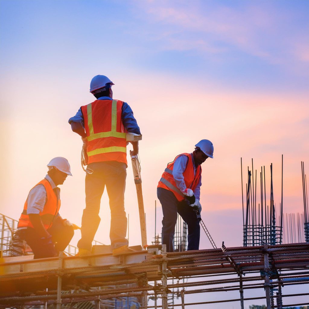 Drei Bauarbeiter tragen Schutzhelme und reflektierende Westen und arbeiten auf einer Baustelle im Freien während des Sonnenuntergangs. Sie stehen auf einem Gerüst oder einer Plattform und führen Bauarbeiten durch. Der Himmel im Hintergrund ist in warmen Farben, was eine stimmungsvolle Atmosphäre schafft.