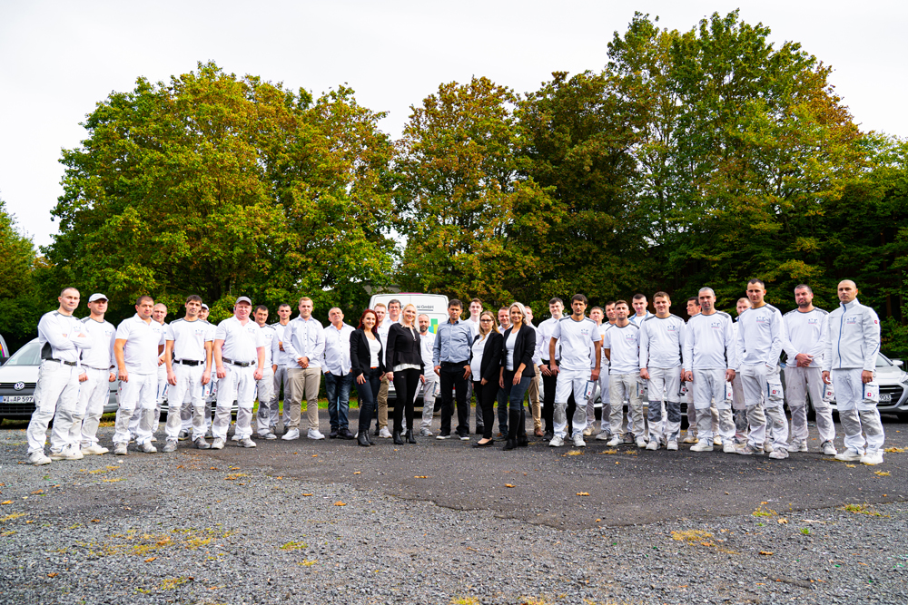 Eine große Gruppe von Menschen, darunter Handwerker in weißen Arbeitsanzügen und Personen in formeller Kleidung, steht draußen auf einem Parkplatz vor einer Reihe von Bäumen. Die Gruppe scheint für ein Gruppenfoto posiert zu haben.