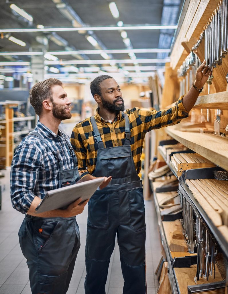 Zwei Handwerker in karierten Hemden und Latzhosen stehen vor einem Regal mit Werkzeugen und Materialien in einem Baumarkt. Einer der Männer zeigt auf ein Werkzeug, während der andere ein Klemmbrett hält und darauf schaut.