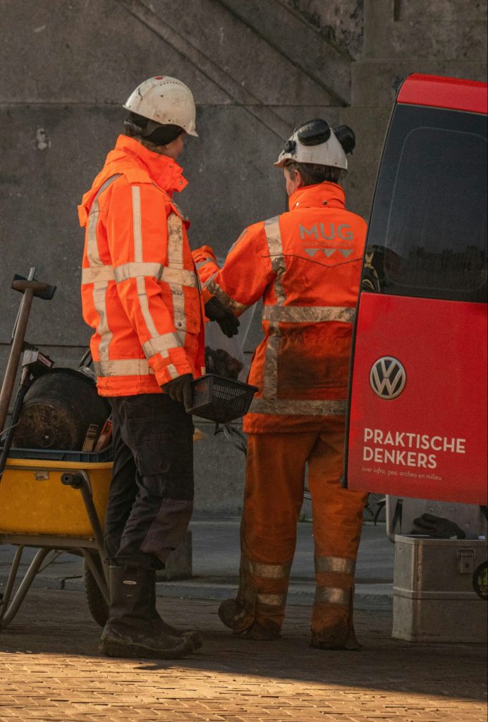 Zwei Bauarbeiter in leuchtend orangefarbenen Schutzjacken und Helmen stehen nebeneinander und unterhalten sich. Sie stehen neben einem roten Lieferwagen mit der Aufschrift „Praktische Denkers“ und einer Schubkarre voller Werkzeuge. Die Szene scheint sich auf einer Baustelle oder einem Straßenbauprojekt abzuspielen.