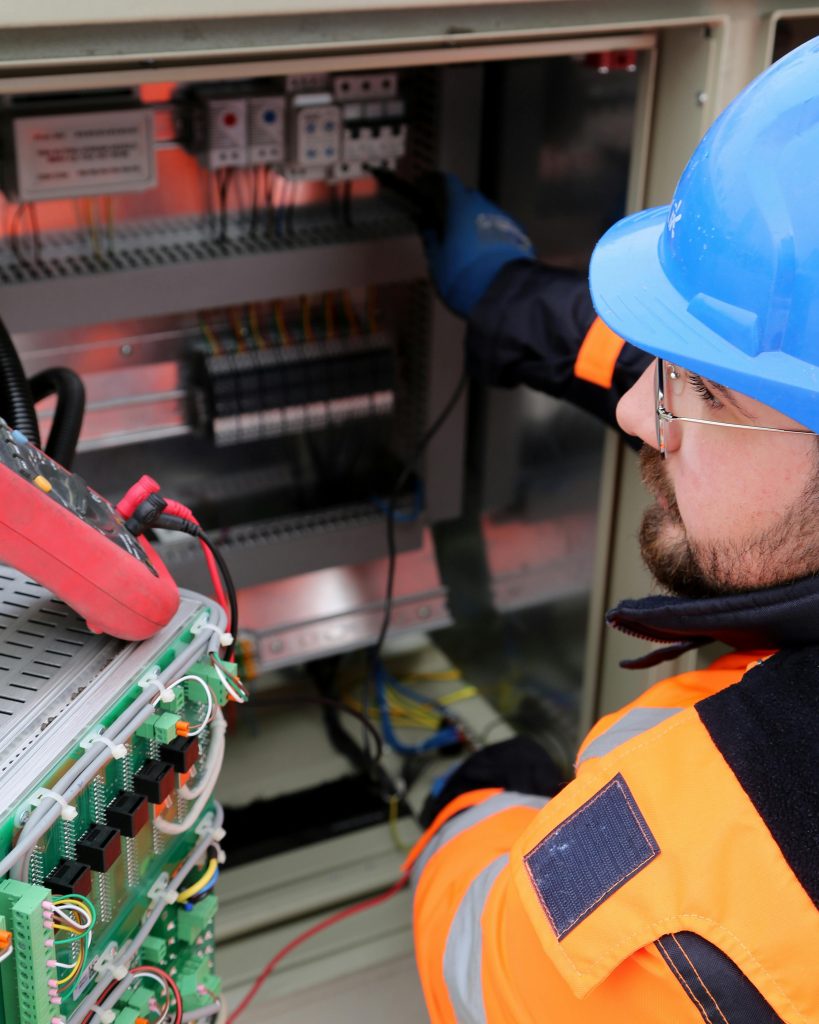 Ein Elektriker mit Schutzhelm und Warnweste arbeitet an einem offenen Schaltschrank, in dem zahlreiche Kabel und Schaltkreise sichtbar sind.