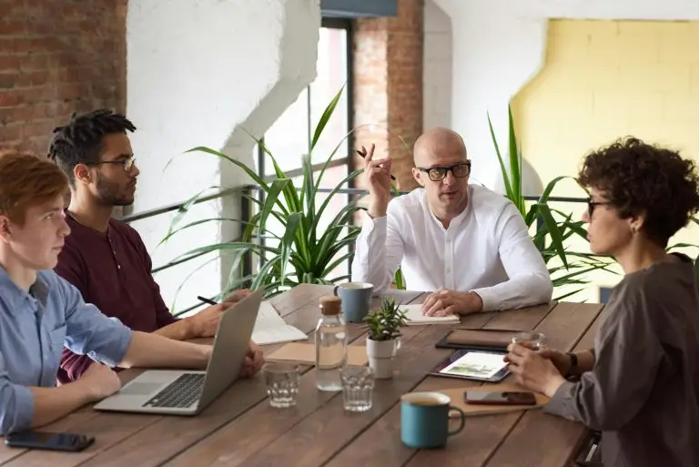 Vier Personen sitzen um einen Tisch in einem modernen Büro und führen ein Meeting. Ein Mann in einem weißen Hemd spricht und gestikuliert, während die anderen aufmerksam zuhören. Auf dem Tisch liegen Laptops, Notizbücher und Kaffeetassen. Im Hintergrund sind Pflanzen und eine Backsteinmauer zu sehen, die dem Raum eine entspannte, aber professionelle Atmosphäre verleihen.