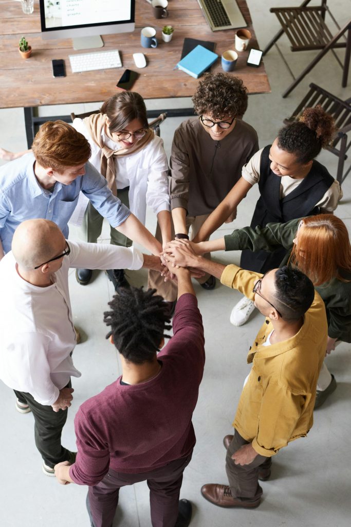 Eine Gruppe von acht Personen steht im Kreis und legt die Hände in der Mitte übereinander, als Zeichen von Teamgeist und Zusammenhalt. Die Szene findet in einem Büro statt, das im Hintergrund teilweise sichtbar ist, mit einem Tisch, Laptops und Kaffeetassen.