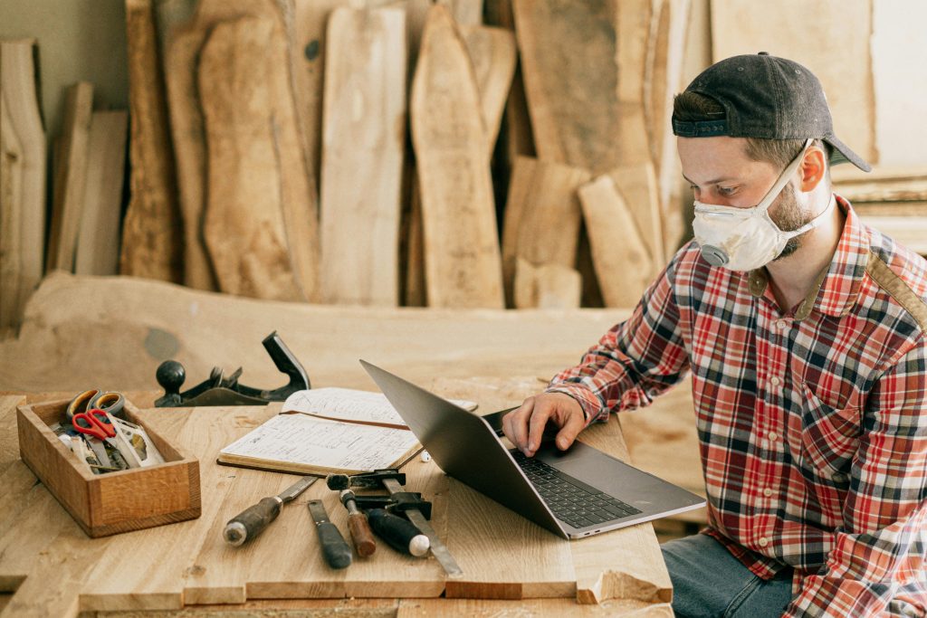 Ein Mann, der eine kariertes Hemd und eine Kappe trägt, arbeitet in einer Werkstatt. Er sitzt an einem Tisch mit verschiedenen Werkzeugen, darunter ein Hammer, Schraubendreher und eine Werkzeugkiste. Der Mann trägt eine Schutzmaske und konzentriert sich auf seinen Laptop, während er in einer handwerklichen Umgebung arbeitet. Im Hintergrund sind große Holzplanken zu sehen.