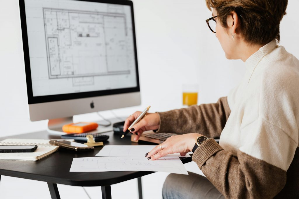 Eine Frau sitzt an einem Schreibtisch und arbeitet an architektonischen Plänen, die auf einem großen Computerbildschirm angezeigt werden. Sie hält einen Stift in der Hand und macht Notizen auf einem Blatt Papier. Auf dem Schreibtisch liegen weitere Büroartikel und ein Glas mit einem Getränk.