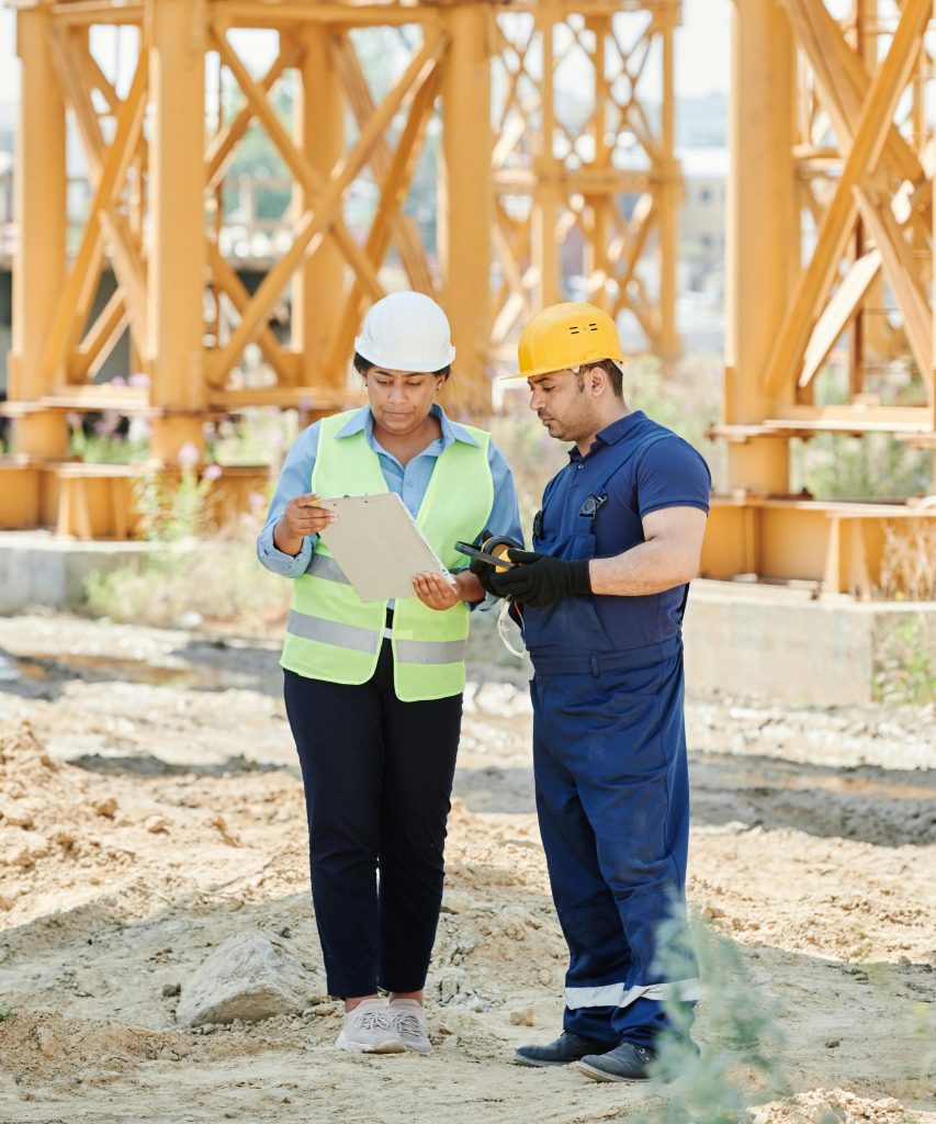 Zwei Bauarbeiter stehen auf einer Baustelle und diskutieren über Pläne. Die Frau links trägt einen weißen Schutzhelm und eine Sicherheitsweste, während sie ein Klemmbrett hält und darauf zeigt. Der Mann rechts trägt einen gelben Schutzhelm, blaue Arbeitskleidung und Handschuhe, und hört der Frau aufmerksam zu. Im Hintergrund sind große Metallstrukturen der Baustelle sichtbar.