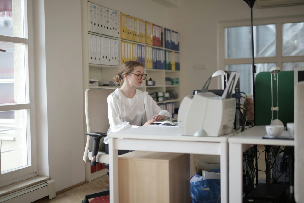 Eine Frau sitzt in einem Büro an einem Schreibtisch, auf dem ein alter Computermonitor steht. Sie ist in weiße Kleidung gekleidet und scheint konzentriert zu arbeiten, während sie in einem Buch oder Dokument blättert. Im Hintergrund befinden sich Regale mit Ordnern in verschiedenen Farben, die ordentlich gestapelt sind.
