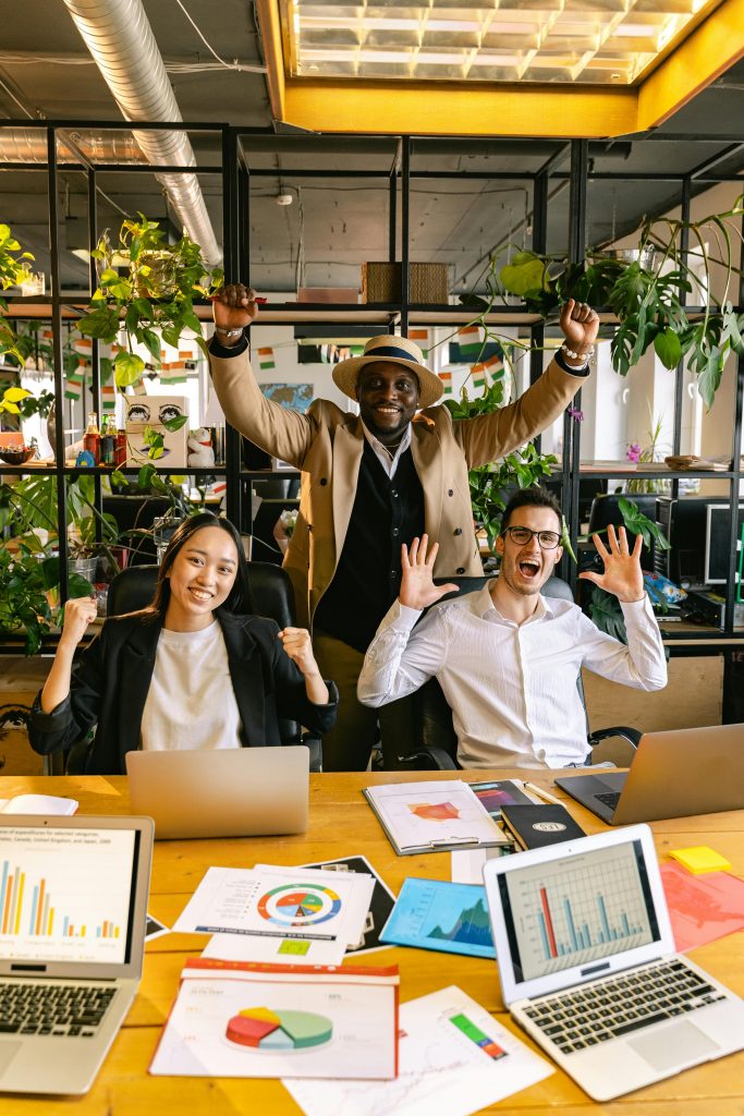 Drei Personen feiern in einem modernen Büro mit einem Pflanzenhintergrund. Zwei von ihnen sitzen an einem Tisch mit Laptops und bunten Diagrammen, die auf dem Tisch verteilt sind, während die dritte Person im Hintergrund steht und beide Arme jubelnd in die Höhe hebt. Alle sind offensichtlich begeistert und feiern ein erfolgreiches Ergebnis.