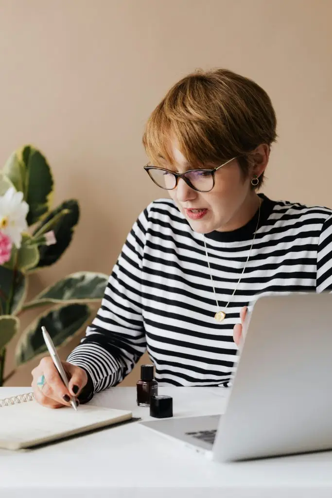 Eine Frau mit kurzen Haaren und Brille sitzt an einem Schreibtisch und arbeitet mit einem Laptop. Sie trägt ein gestreiftes Oberteil und hält einen Stift in der Hand, während sie in ein Notizbuch schreibt. Auf dem Tisch stehen auch einige Tintenflaschen und ein weiteres Notizbuch. Im Hintergrund ist eine Pflanze sichtbar, die dem Raum eine angenehme Atmosphäre verleiht.