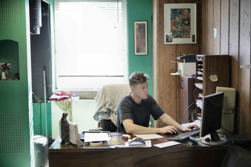 Ein junger Mann sitzt in einem kleinen, leicht beengten Büro und arbeitet an einem Computer. Der Raum hat grüne und holzgetäfelte Wände, mit einem Fenster im Hintergrund, das Tageslicht hereinlässt. Auf dem Schreibtisch liegen verschiedene Papiere und Büroartikel, und an der Wand hängt ein gerahmtes Bild. Der Mann scheint konzentriert zu arbeiten.