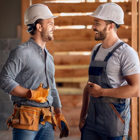 Zwei Bauarbeiter stehen auf einer Baustelle und unterhalten sich fröhlich. Beide tragen weiße Schutzhelme. Der Arbeiter links trägt ein kariertes Hemd, Jeans und einen Werkzeuggürtel, während der Arbeiter rechts in einer grauen Latzhose und einem T-Shirt gekleidet ist. Sie lachen und wirken zufrieden, während sie sich in einer halbgebauten Holzkonstruktion unterhalten.