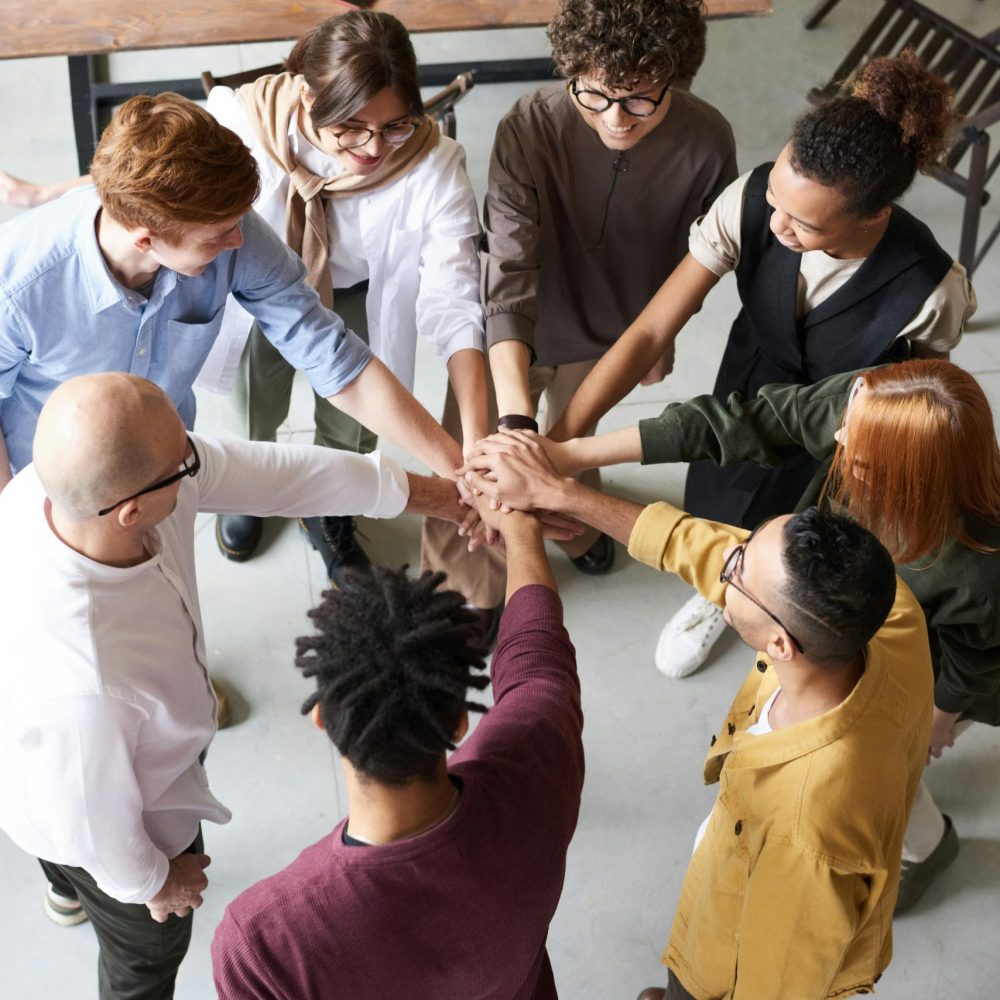 Eine Gruppe von acht Personen steht im Kreis und legt die Hände in der Mitte übereinander, als Zeichen von Teamgeist und Zusammenhalt. Die Szene findet in einem Büro statt, das im Hintergrund teilweise sichtbar ist, mit einem Tisch, Laptops und Kaffeetassen.